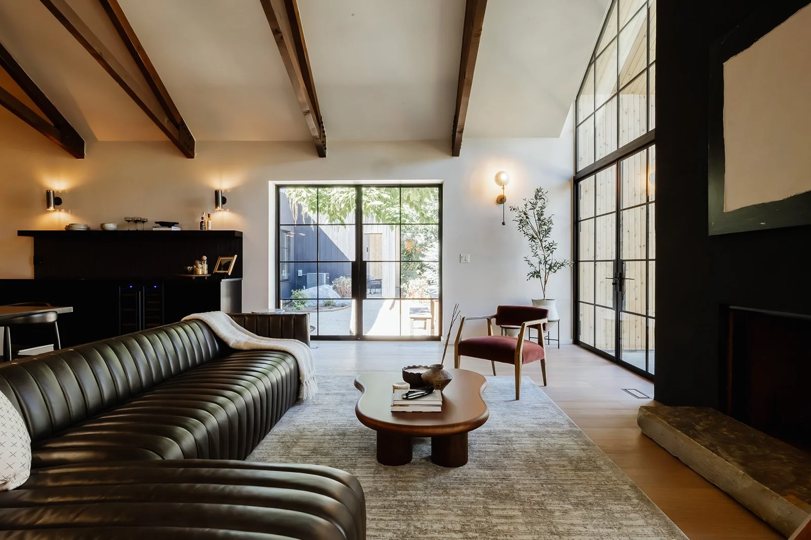 Modern living room with black leather couch, wooden coffee table, armchair, large windows, and minimal decor.