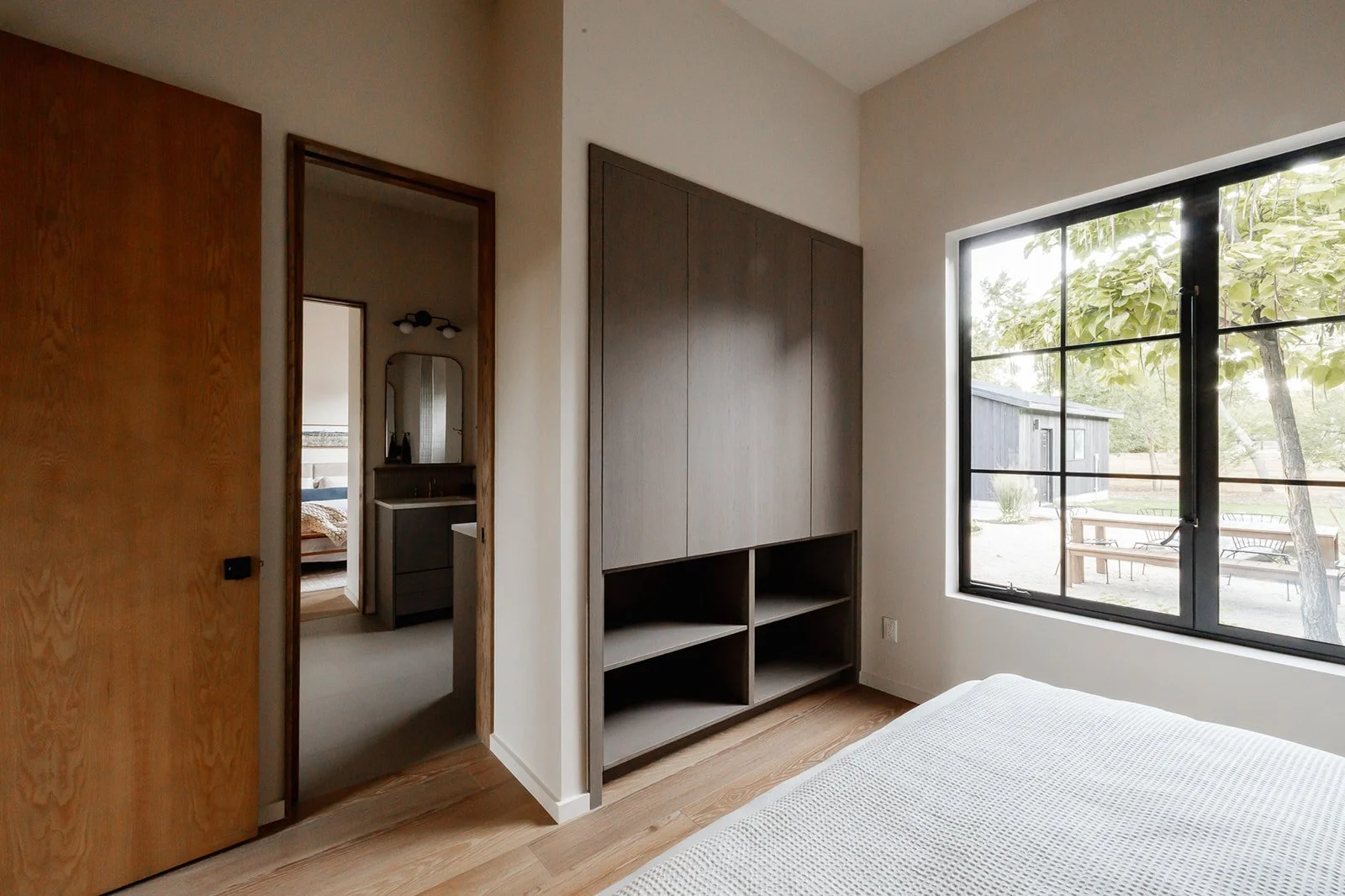 A bedroom with a large window showing an outdoor area, a bed with light-colored bedding, a tall cabinet, and a doorway leading to a bathroom with a mirror and sink.