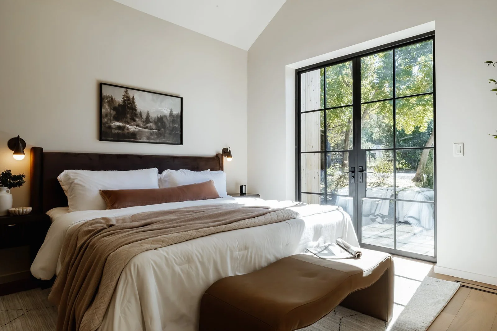 Bright bedroom with a large bed, dark wood headboard, white bedding, tan blanket, and a brown pillow. There are wall-mounted reading lights, a bedside table with a plant, and a large black-framed glass door leading to a patio with greenery outside.