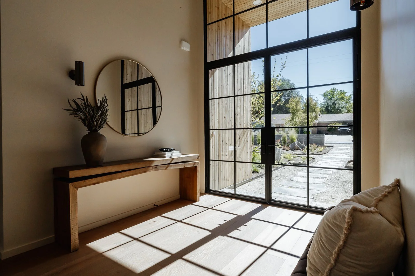 Sunlit modern entryway with large glass door, wooden accents, a bench, a round mirror, and a decorative vase with plants.