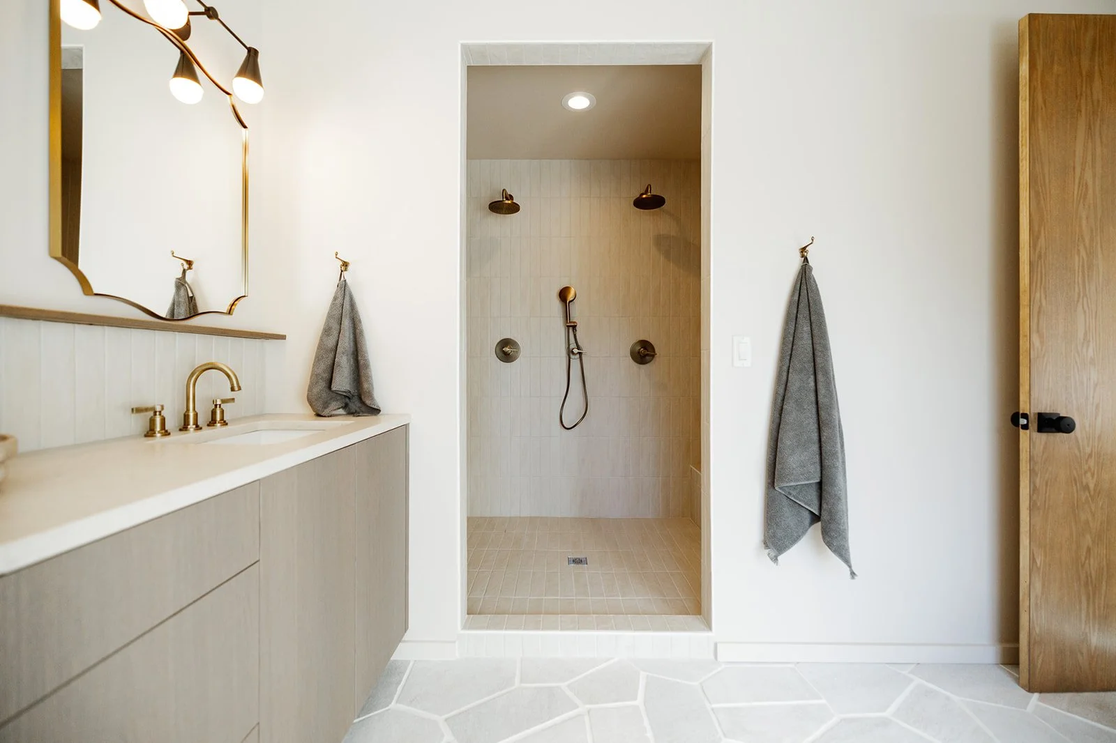Modern bathroom with a vanity and mirror on the left, an open shower with two showerheads in the center, and a wooden cabinet on the right. Gray towels hang on hooks on the wall.