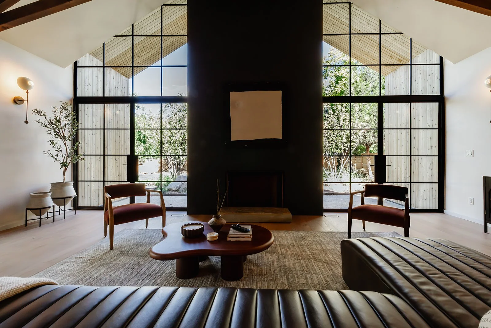 Living room with large windows, black framed glass, a fireplace with a white artwork above, two brown armchairs, a wooden coffee table, a striped leather sofa, and indoor plants.