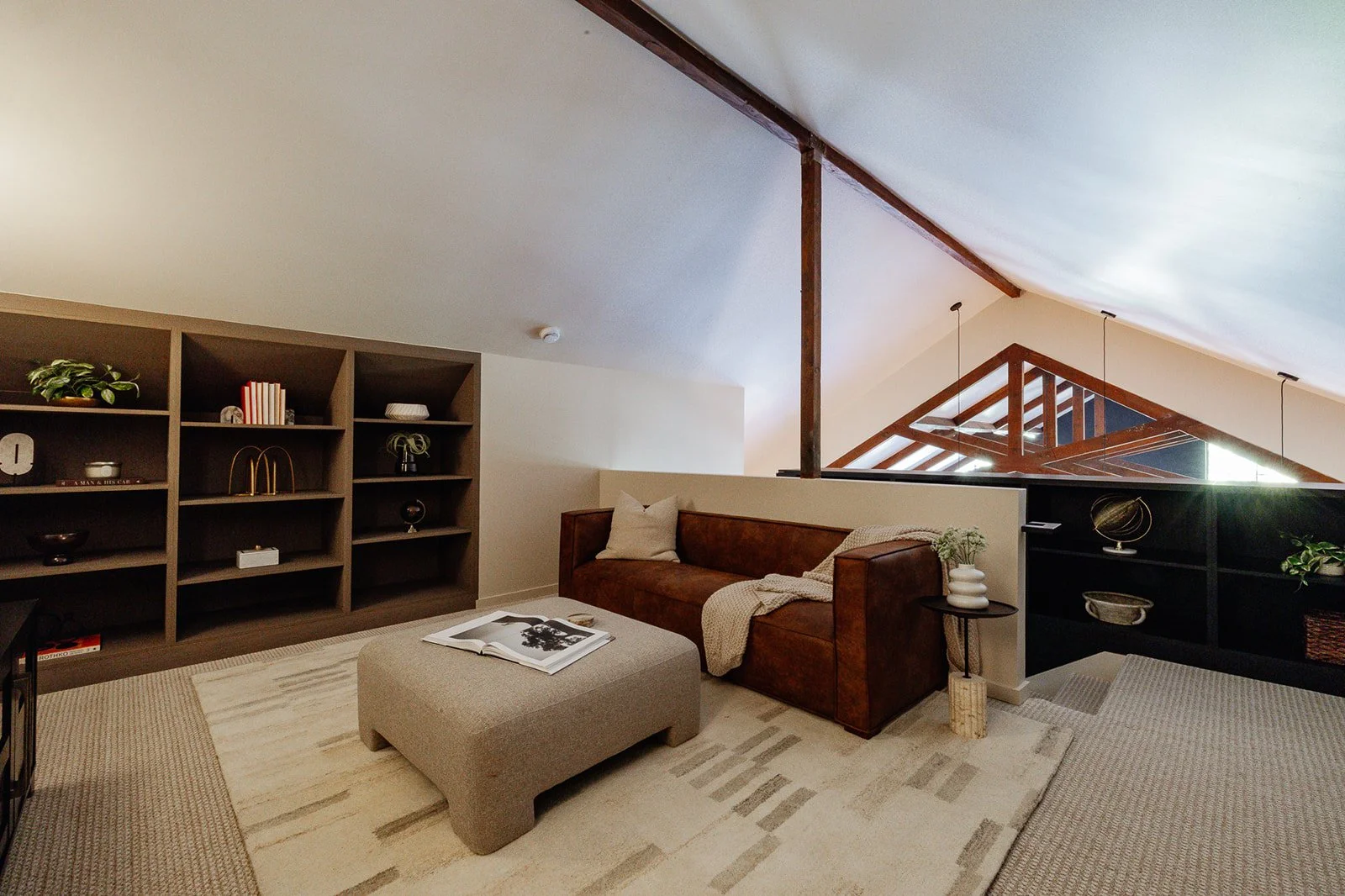 Living room with a brown leather sofa, an ottoman with an open magazine, a cream-colored area rug, a black bookshelf, and a side table with a white vase and greenery. Exposed wooden beams are visible in the ceiling.