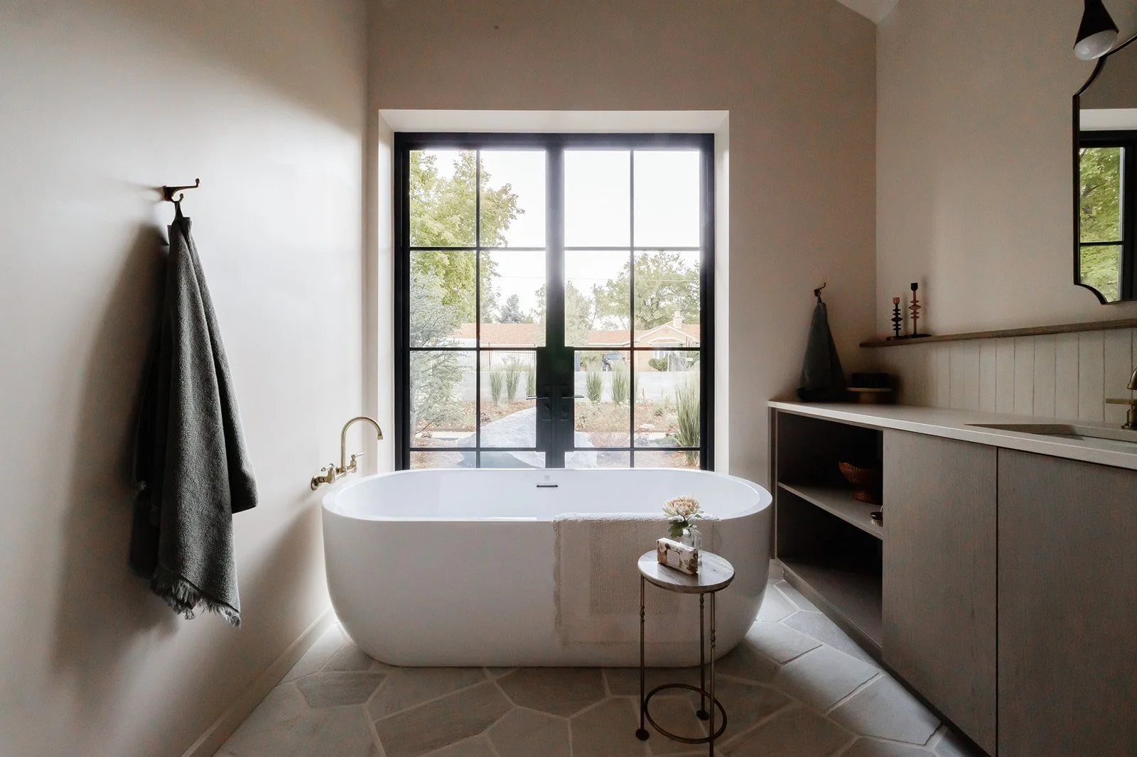 Modern bathroom with a large bathtub in front of a window, a gray towel hanging on a hook on the wall, a small side table with a flower and toiletries, and a gray vanity with a mirror and decorative items.