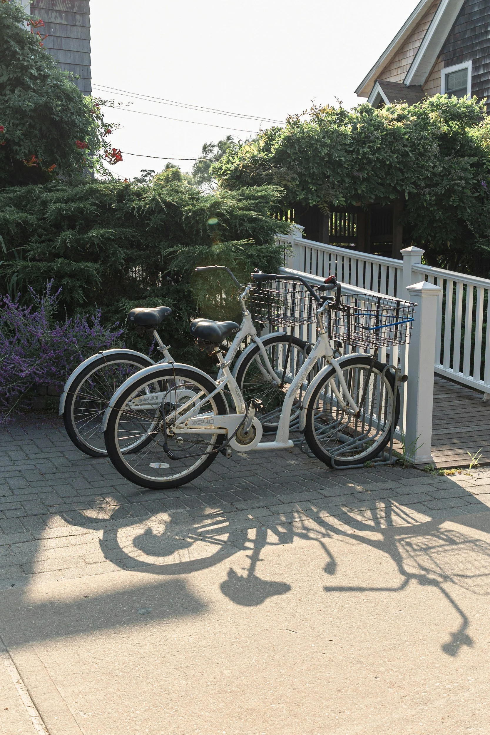 Two white bicycles with black seats and a front basket parked side by side on a brick-paved sidewalk near a white wooden railing. The bicycles cast shadows on the ground, and there are green bushes and a house in the background.