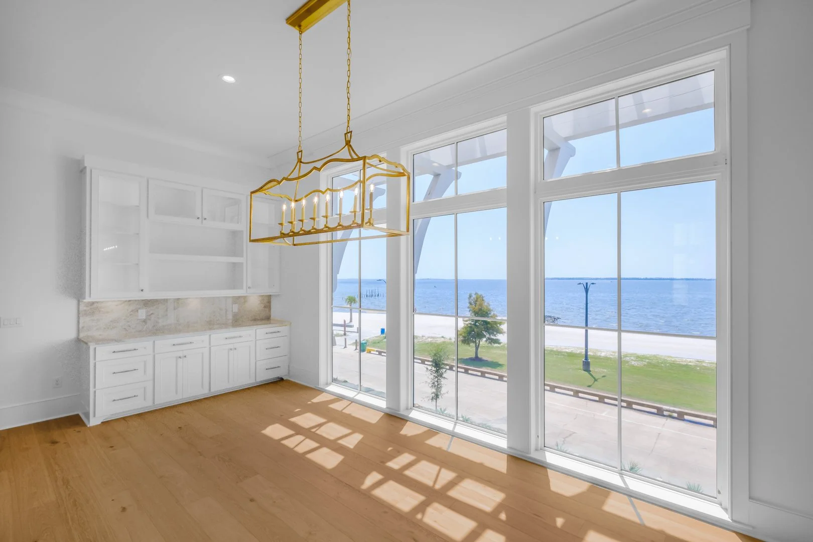 Empty dining area with large windows overlooking ocean, white cabinetry, wood floor, and gold chandelier.