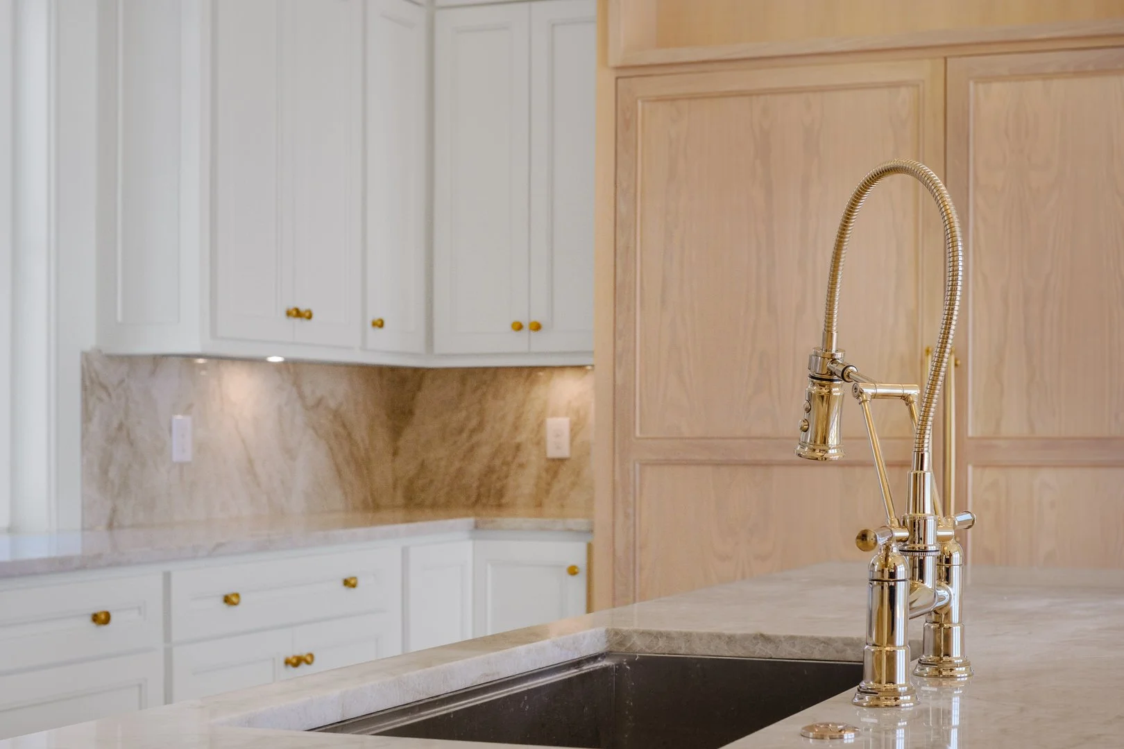 Kitchen sink with a gold faucet on a marble countertop, wooden and white cabinets with gold knobs in the background.