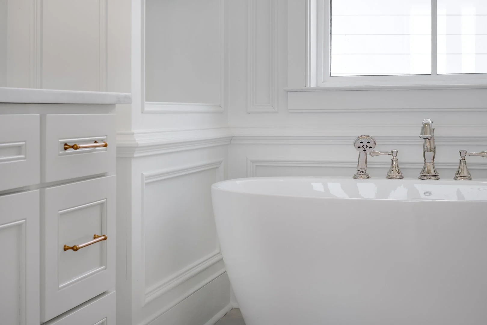 A white bathroom with a freestanding bathtub near a window and white cabinetry with wooden handles.