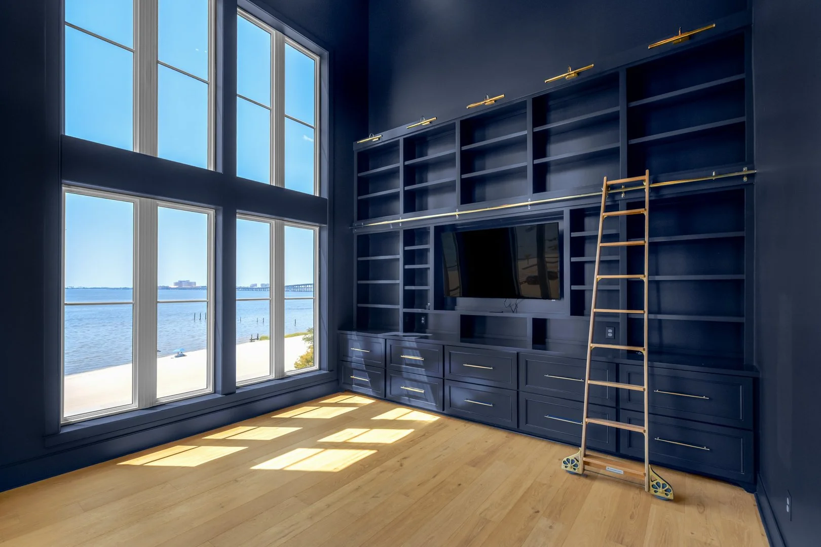 Empty room with large windows overlooking water, blue walls, and built-in dark blue wall unit with a television, shelving, and drawers, with a wooden ladder leaning against the unit.