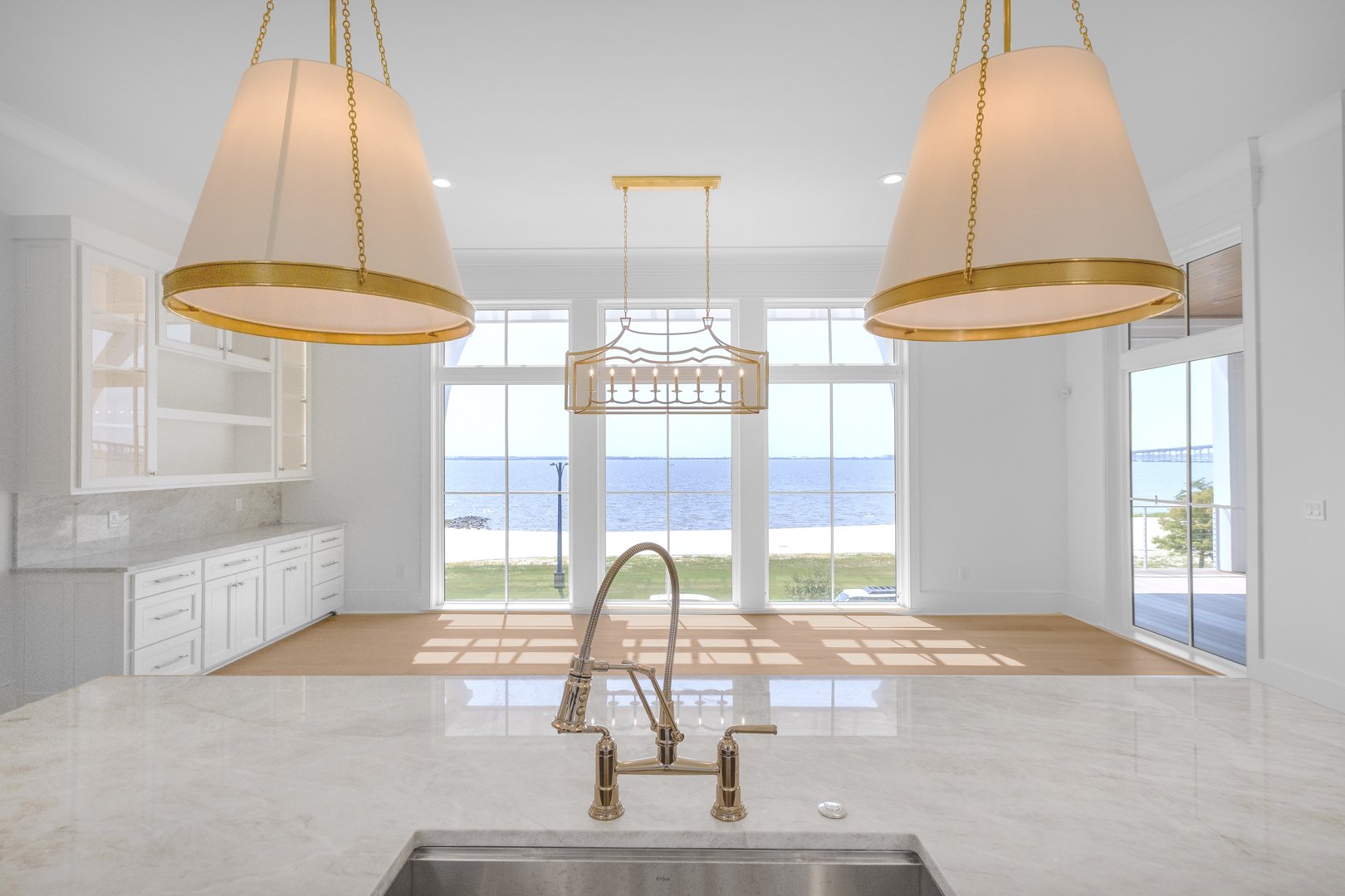 Interior view of a bright kitchen and dining area overlooking a body of water, with large windows, a marble kitchen island with a sink, and white cabinetry.