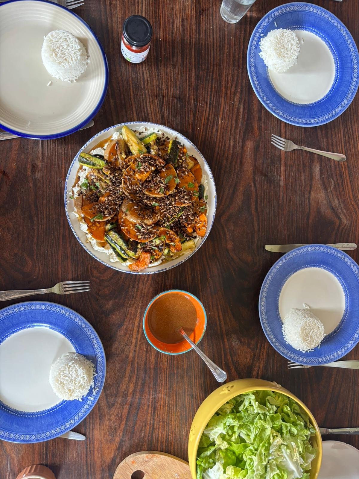 Table avec saladier de légumes sautés, assiettes avec boule de riz, verres d'eau, sauce rouge, une salade verte en plastique jaune, assiettes bleues avec de la crème et du riz, cuisine ayurvédique au domaine de Maynard, gite de groupe.