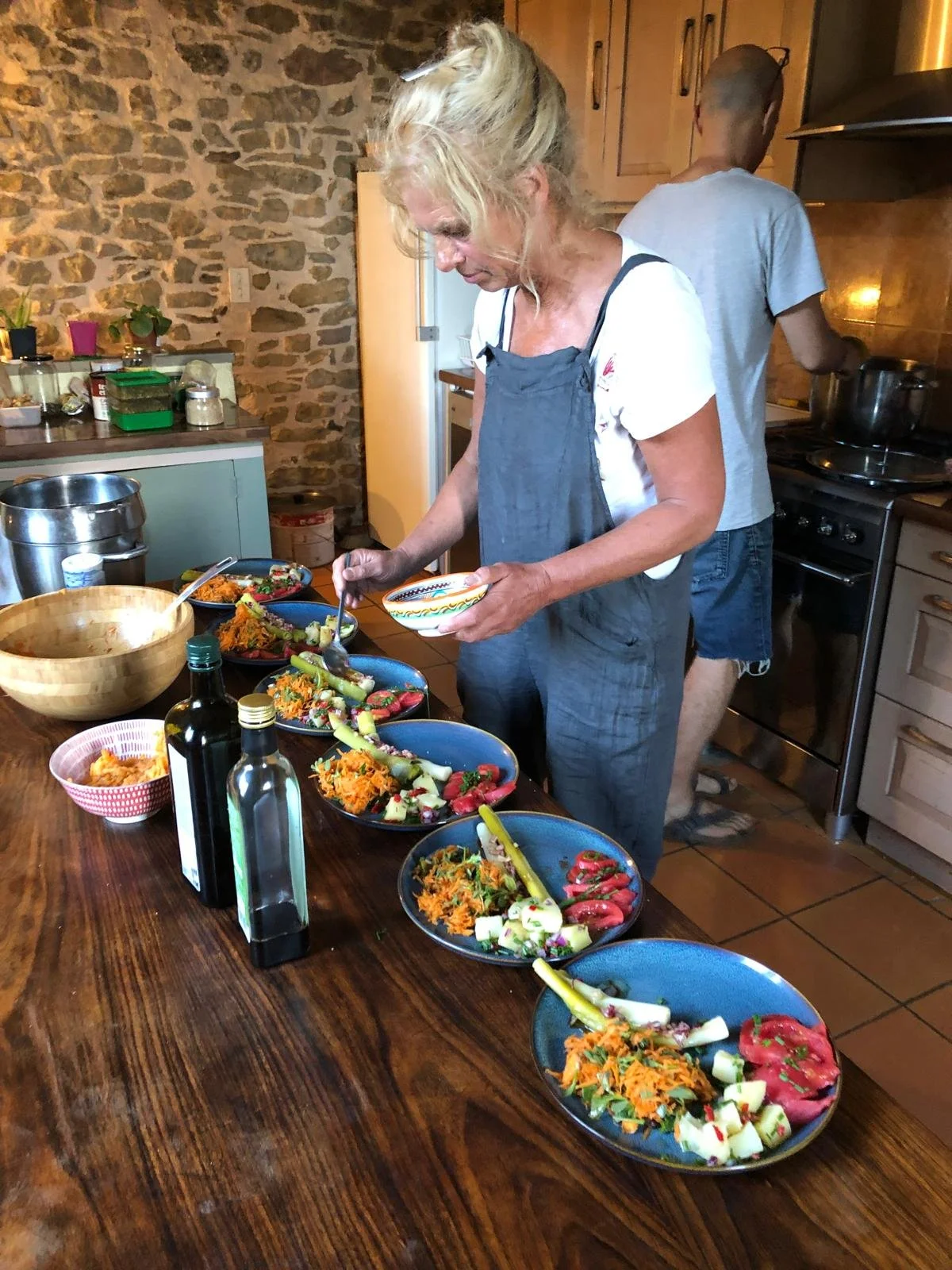 Une femme prépare et sert des plats de nourriture végétarienne ou végétalienne dans une cuisine rustique avec un mur en pierre au Domaine de Maynard, gite de groupe à proximité de Limoux, Toulouse et Carcassone.