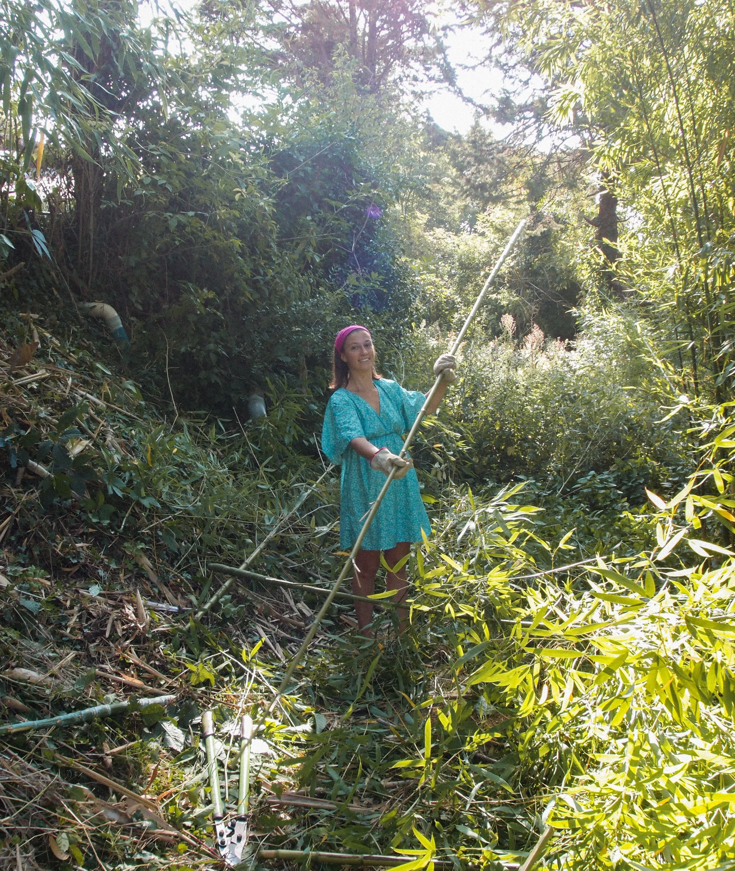 Une femme portant une robe bleue et un bandeau rose, tenant une canne à pêche dans une forêt dense contre la lumière du soleil au Domaine de Maynard, gite de groupe à proximité de Limoux.