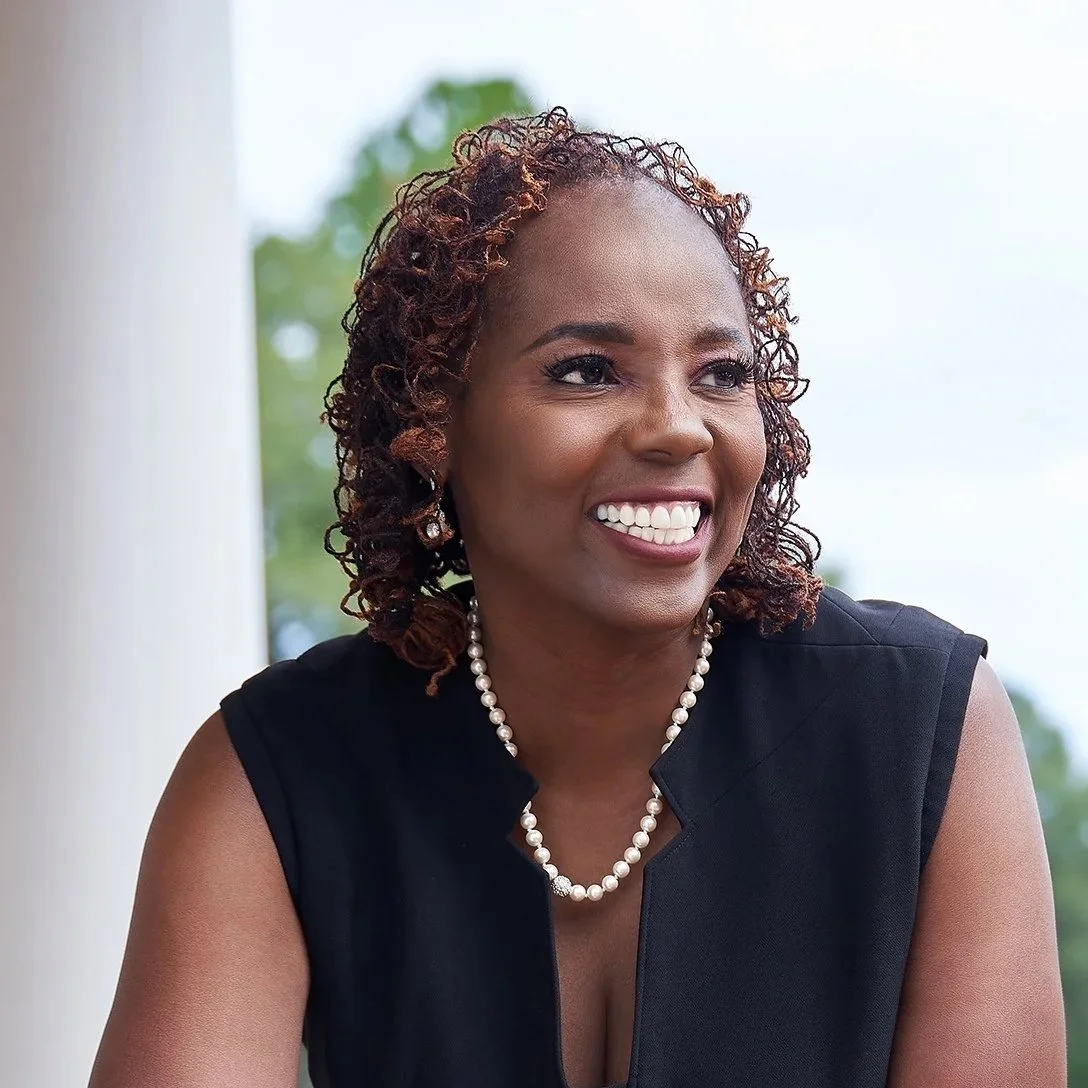 A woman with curly hair, wearing a black sleeveless top, pearl necklace, and earrings, smiling outdoors with greenery in the background.