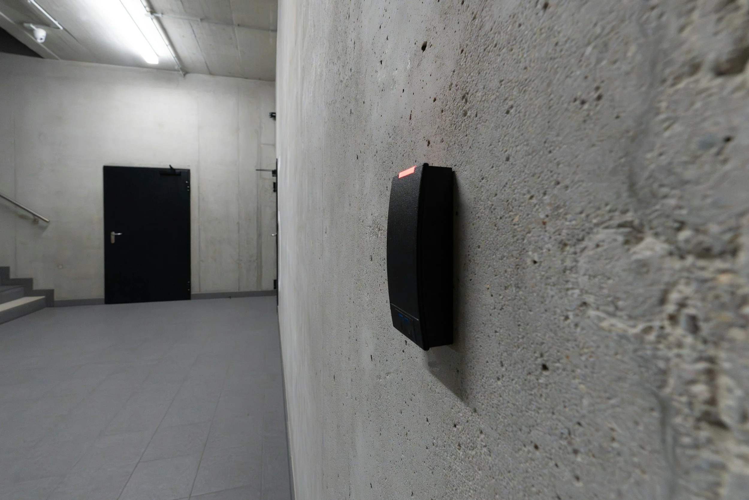 A black card reader placed on a rough concrete wall in a minimalist, industrial-style concrete room with a black door and gray stairs.