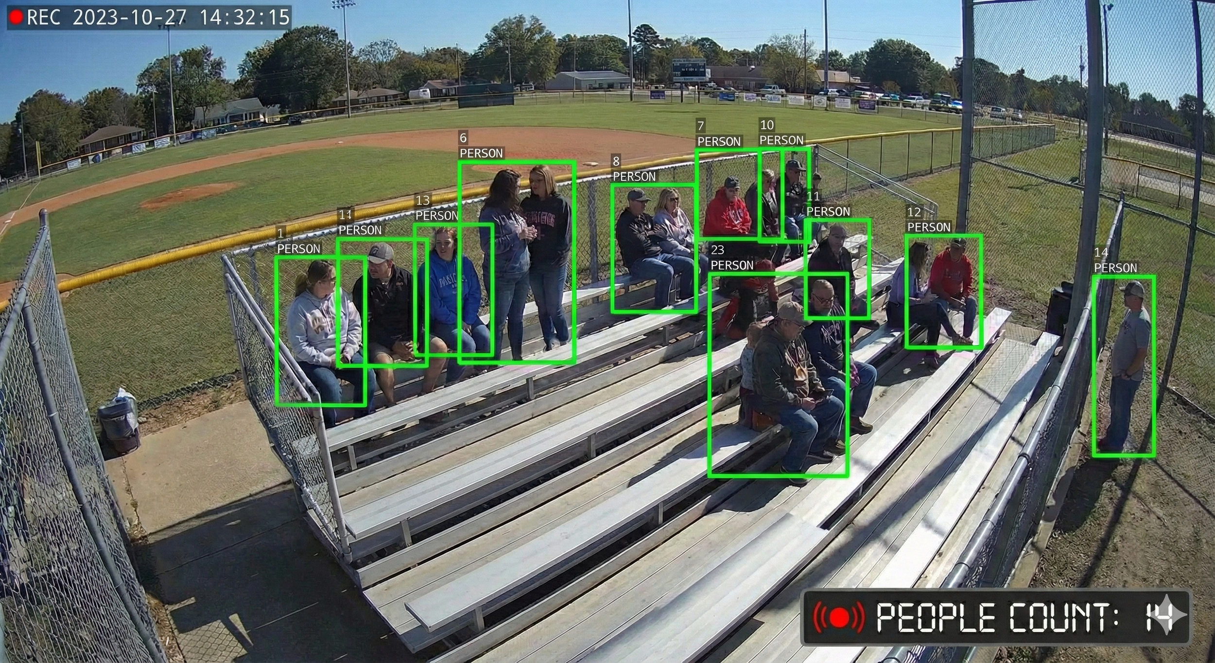 People sitting and standing on bleachers at a baseball field, with some engaged in conversation. The scene is outdoors on a sunny day, surrounded by a chain-link fence and grassy areas.