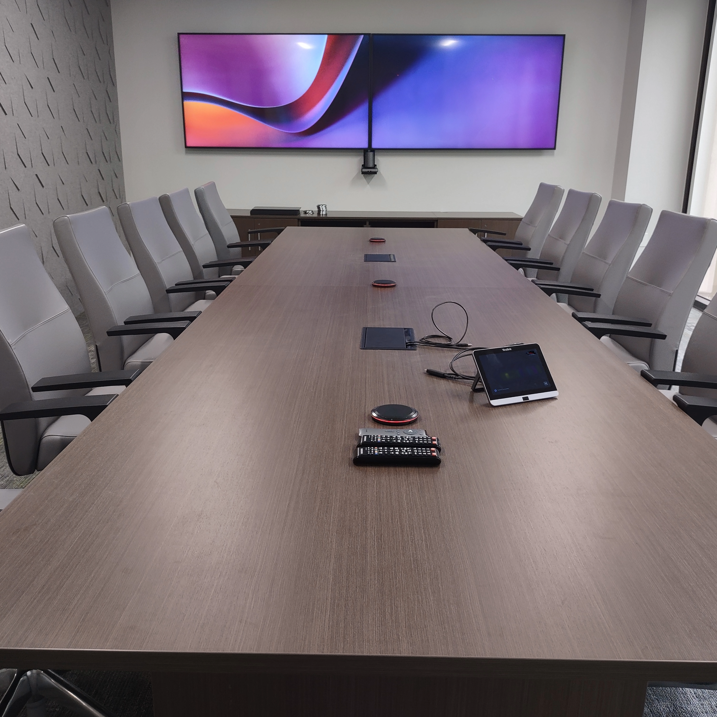 Empty conference room with a large wooden table surrounded by grey office chairs, a wall-mounted screen at the end of the room, and various electronic devices placed on the table.