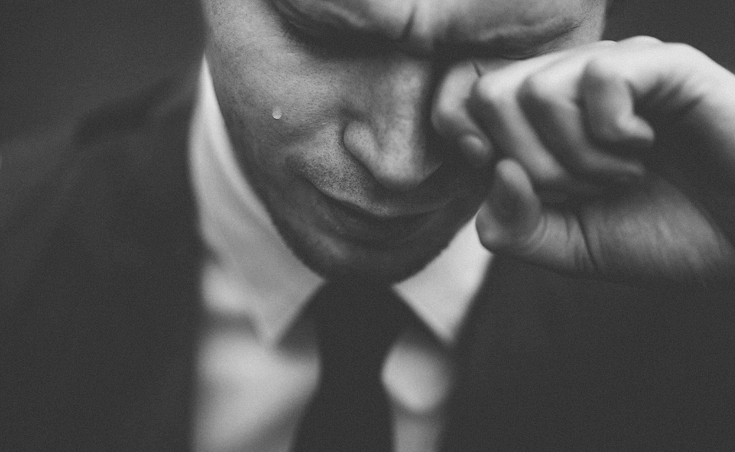 A black and white close-up photograph of a man in a suit, touching his temple with his hand, with a somber expression.