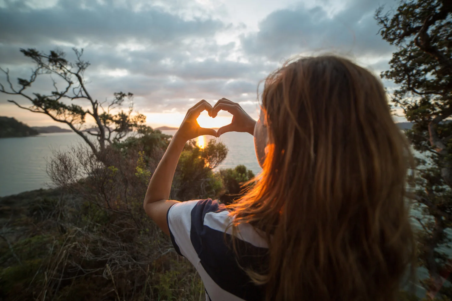 A woman with brown hair making a heart shape with her hands, framing the setting sun over a body of water at sunset, surrounded by trees and clouds.