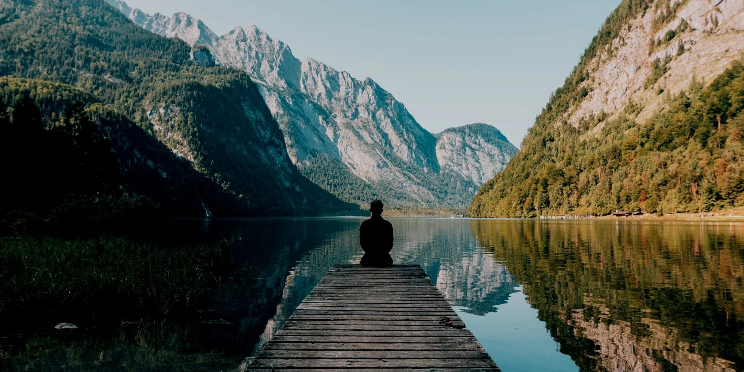 Person sitting on a dock by a calm lake in a mountain valley, with lush green forests and rugged mountains in the background.