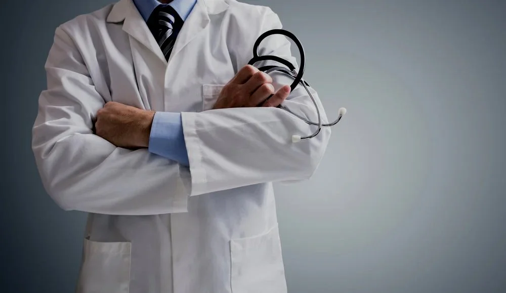 A person in a white doctor's coat holding a stethoscope. The person is wearing a blue shirt and a dark striped tie. Their head is not visible.