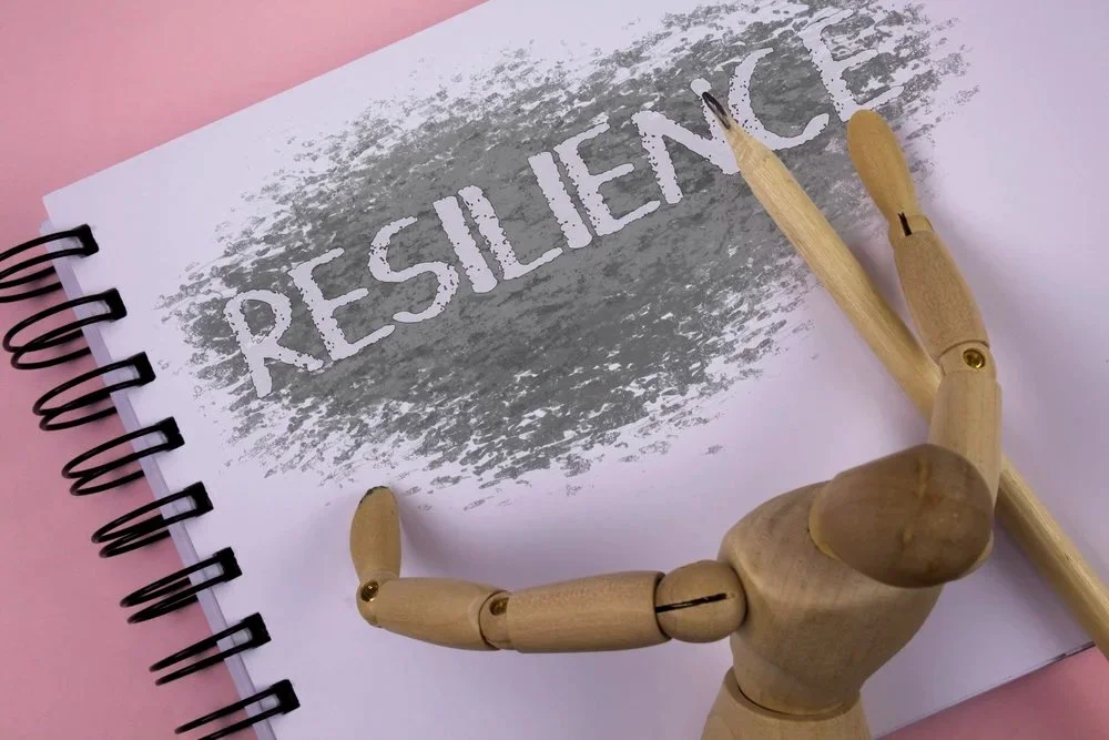 A wooden artist mannequin holding a wooden pen on a white spiral notebook with the word RESILIENCE written in gray paint.