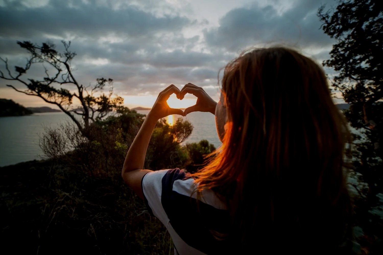 A woman with brown hair making a heart shape with her hands, framing the setting sun over a body of water at sunset, surrounded by trees and cloudy sky.