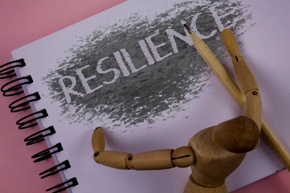 A spiral-bound notebook with the word RESILIENCE in white letters on a gray textured background. A wooden artist mannequin is holding a wooden pencil next to the notebook.