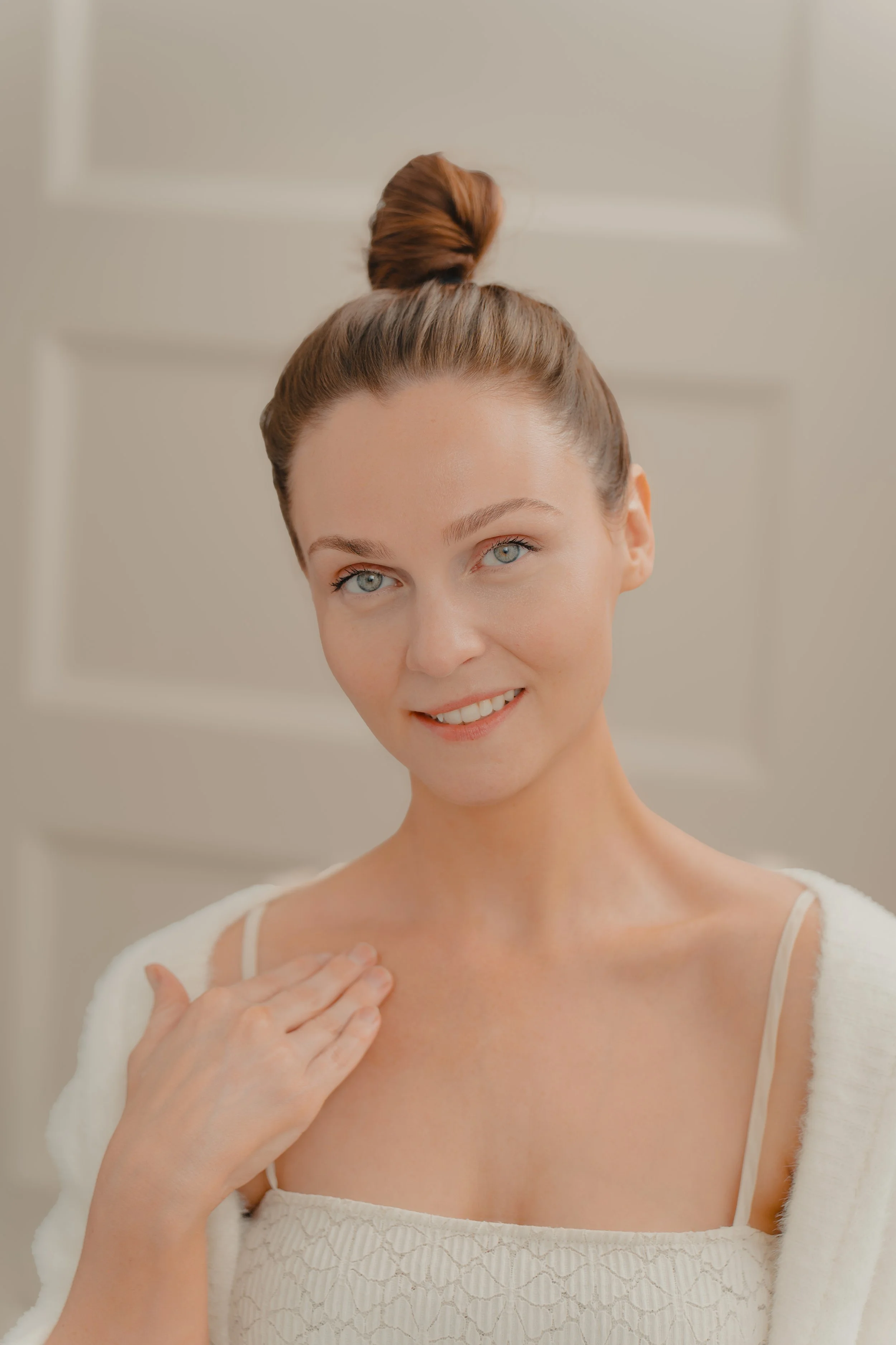 A woman with blue eyes and brown hair in a top bun, smiling at the camera, wearing a cream-colored dress and a white cardigan, in a neutral background.
