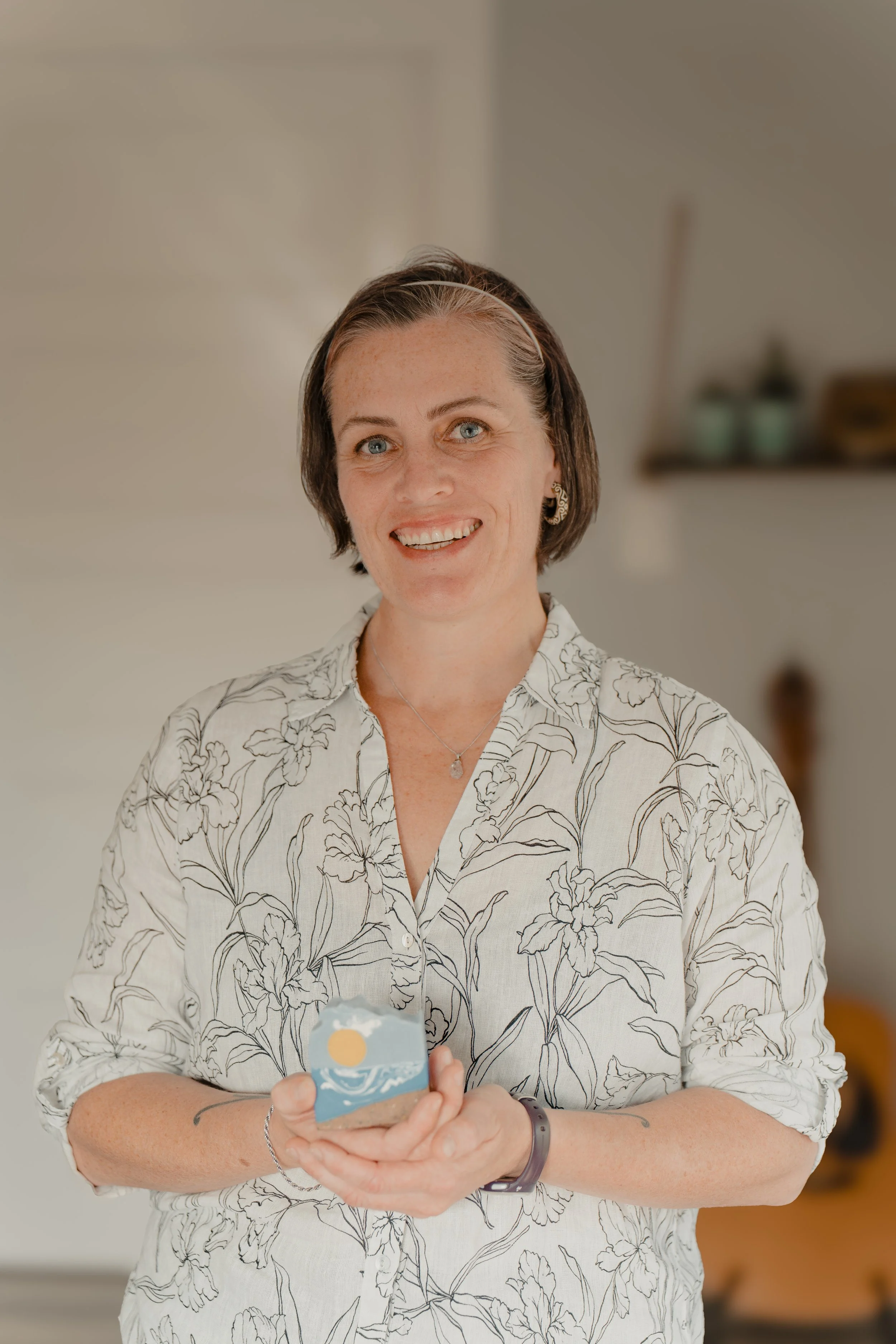 A smiling woman with short brown hair, wearing a floral button-up shirt, holding a small painted rock with a sun and waves design, standing in a cozy, softly-lit room.