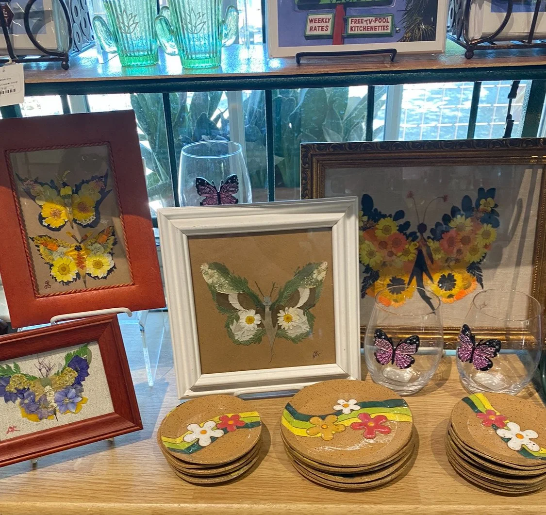 Display of butterfly-themed art, decorative plates, and glassware with butterfly stickers on a wooden table at a shop.