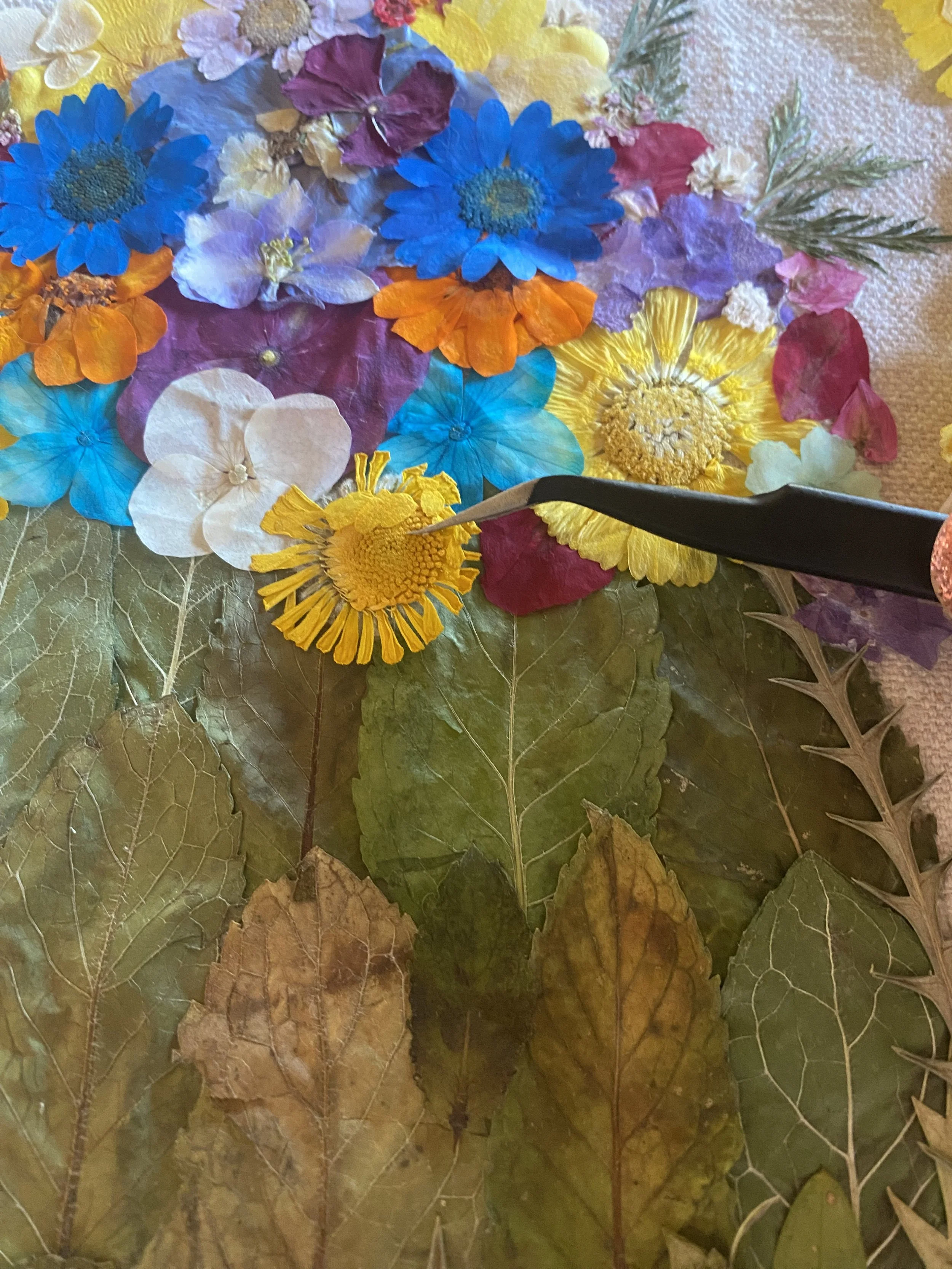 A variety of colorful pressed flowers and leaves arranged on a base, with a tool working on the flowers.