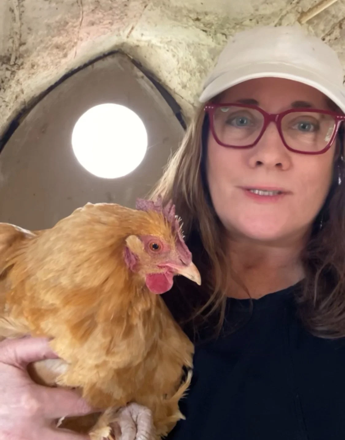 A woman with red glasses and a white cap holding a chicken inside a stone structure with a small round window in the background.