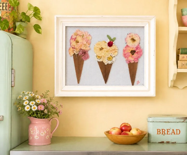 Decorative artwork of three ice cream cones with flower scoops and a cherry, framed on a yellow wall in a cozy kitchen corner.
