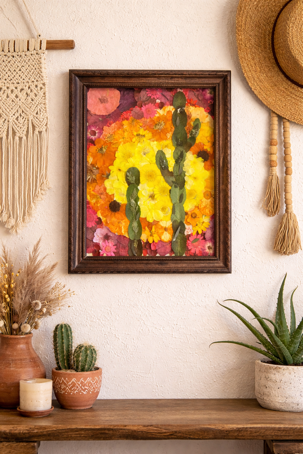Decorative wall art featuring a cactus made of pressed flowers and leaves, framed and hanging on a textured cream wall with plants and woven decorations nearby.