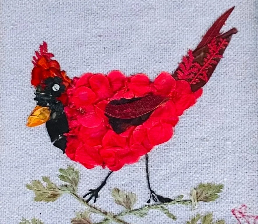 Bird made from red, black, yellow, and brown flower petals and leaves on a light fabric background.