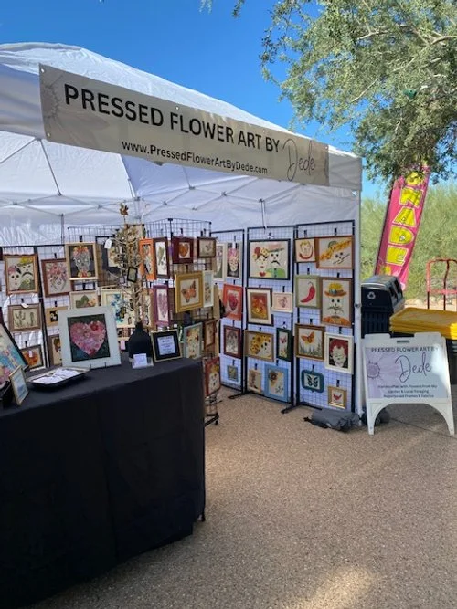 Outdoor art booth showcasing pressed flower artwork with framed pieces of various designs including hearts and animals, under a white canopy labeled "Pressed Flower Art by Dede."