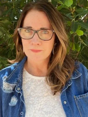 A woman with long wavy brown hair wearing glasses, a white lace top, and a denim jacket outdoors with green foliage in the background.