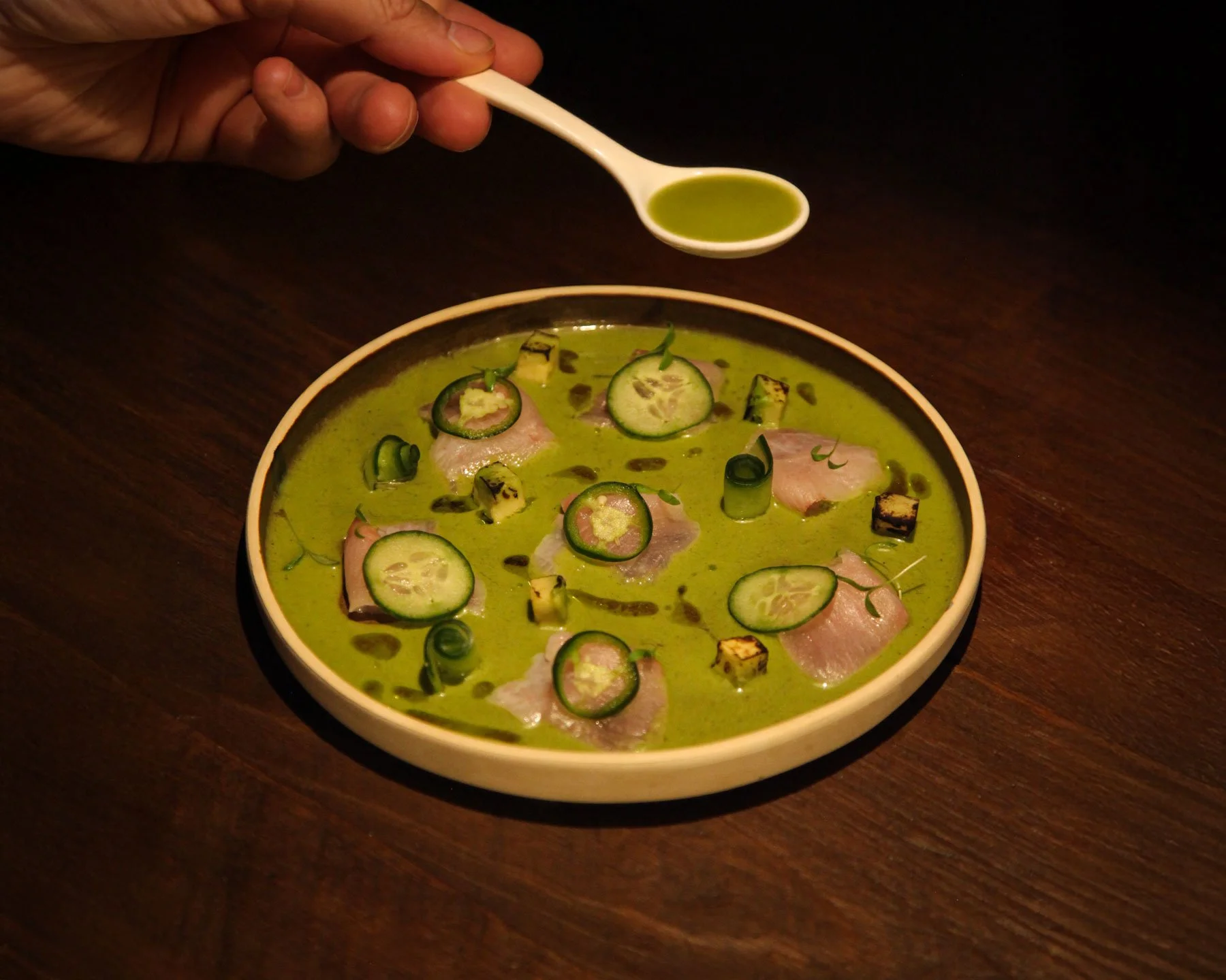 A bowl of green soup garnished with cucumber slices, small cubes of grilled zucchini, and slices of meat, with a small spoon of green sauce being drizzled over it.