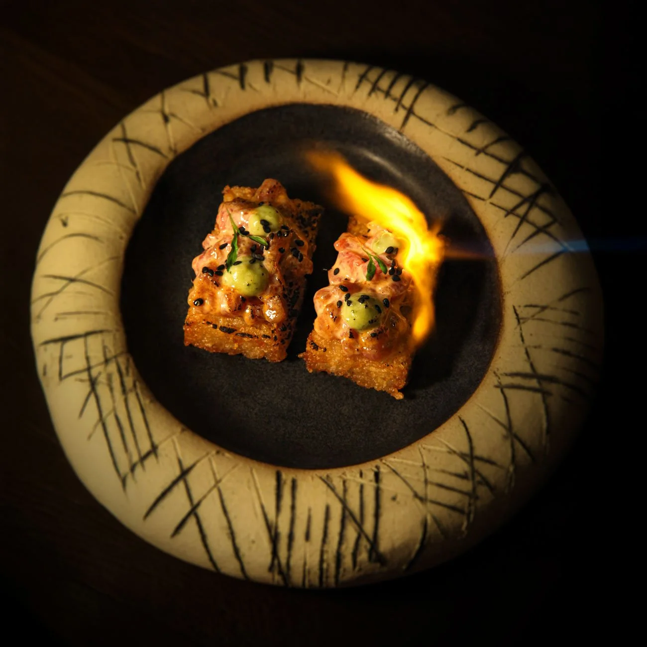 Two pieces of sushi topped with green sauce and black sesame seeds served on a black plate, with a small flame next to them.
