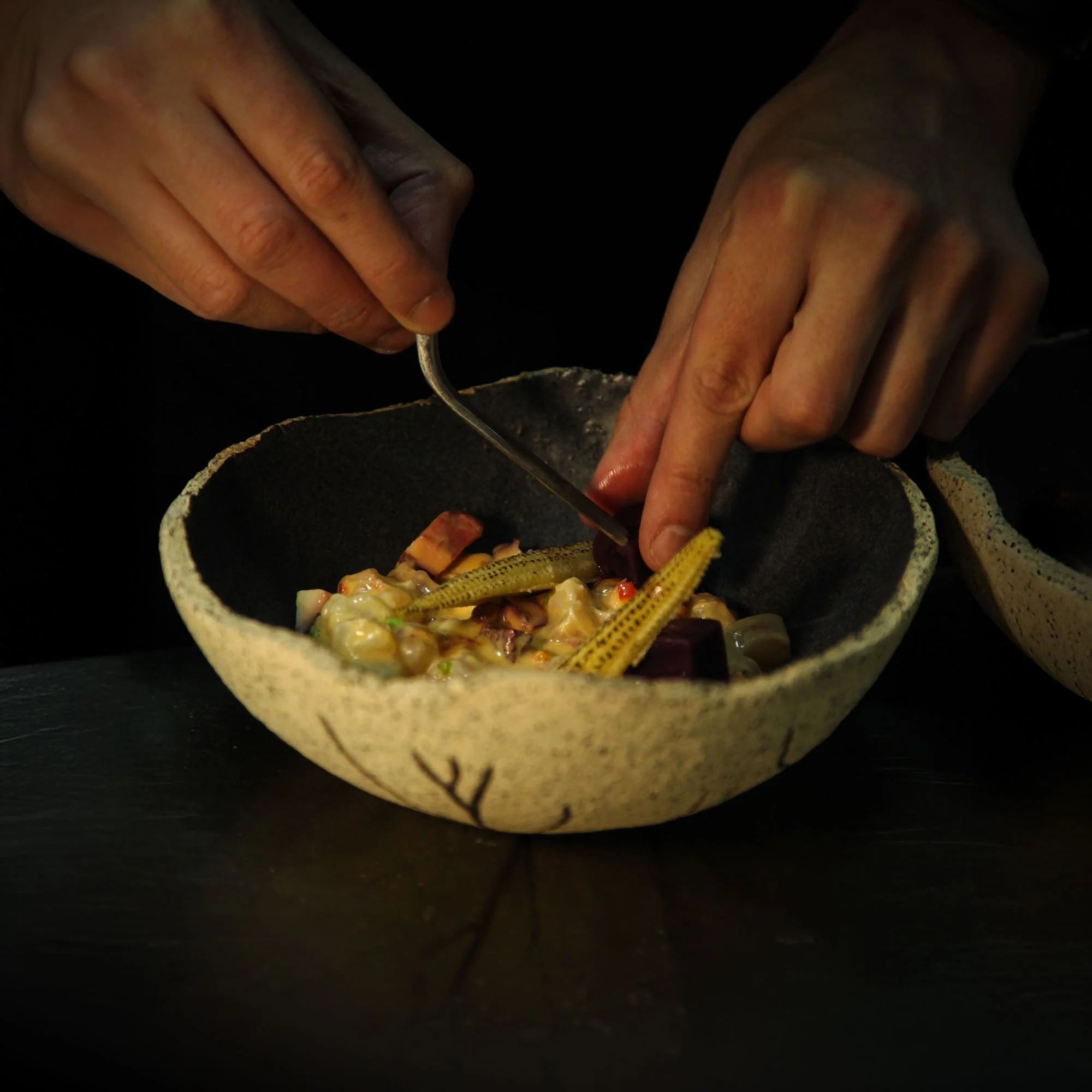Close-up of hands using a fork to serve a colorful dish with vegetables in a rustic bowl.