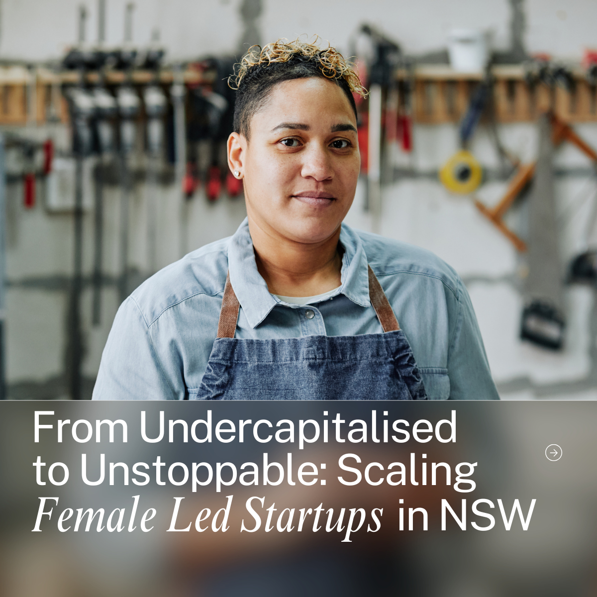 A woman with short curly hair and earrings wearing a denim shirt and apron in a workshop with tools on the wall.