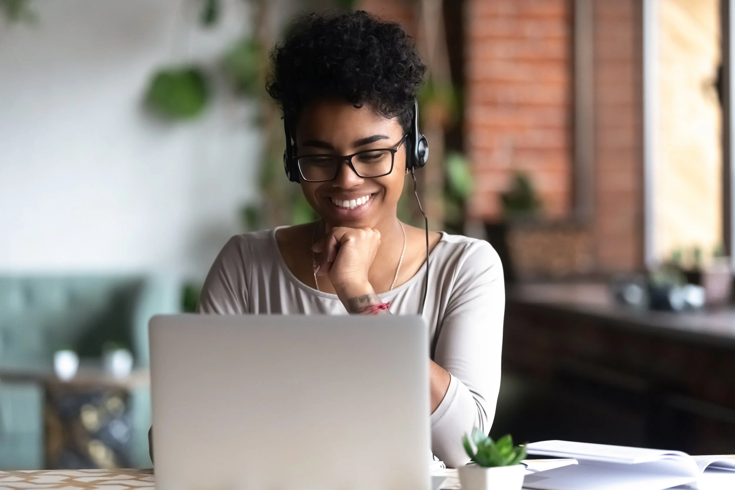 A woman with glasses and curly hair wearing headphones, smiling while looking at her laptop, sitting at a table with a small plant and an open book in a cozy indoor setting.