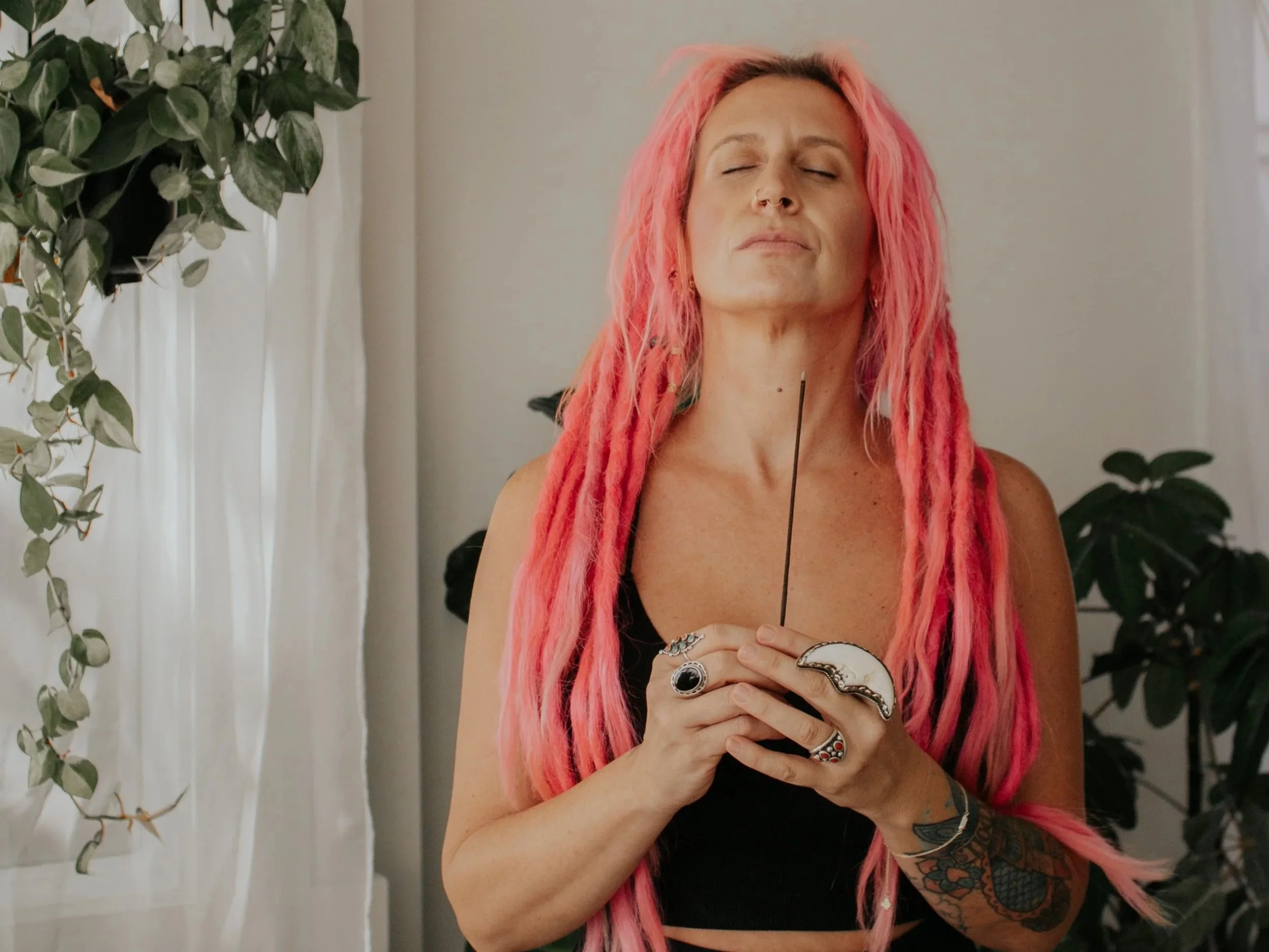 A woman with long pink hair, closed eyes, and a peaceful expression, holding a necklace in her hands, standing indoors with plants and curtains in the background.