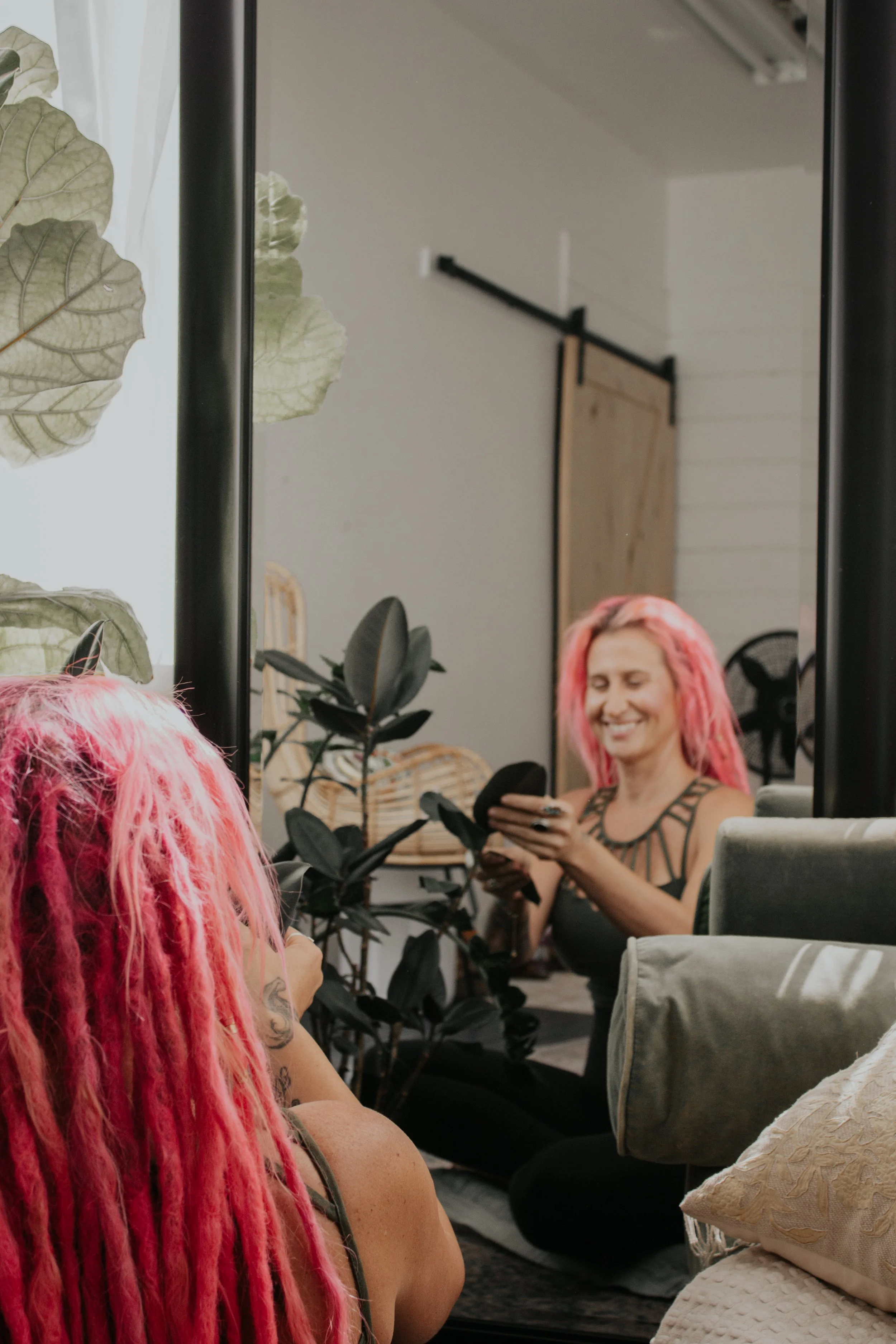 A woman with pink dreadlocks smiling while looking at her phone, with plants and a mirror reflecting her in a cozy indoor space.