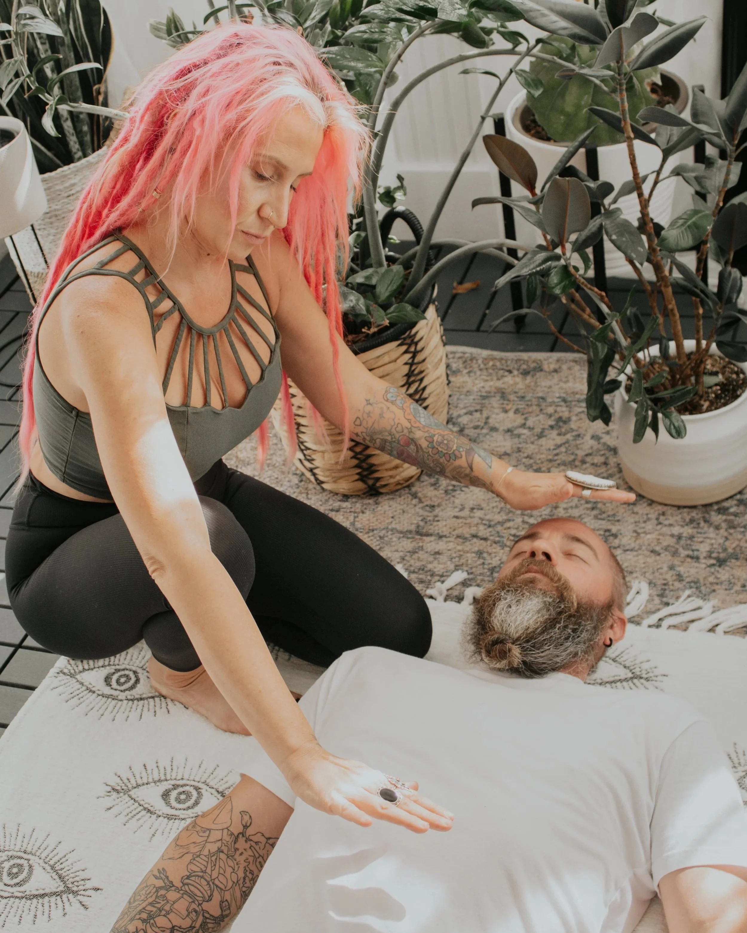 A woman with pink dreadlocks and tattoos is performing a healing or reiki session on a man lying on his back with eyes closed in a cozy, plant-filled space.