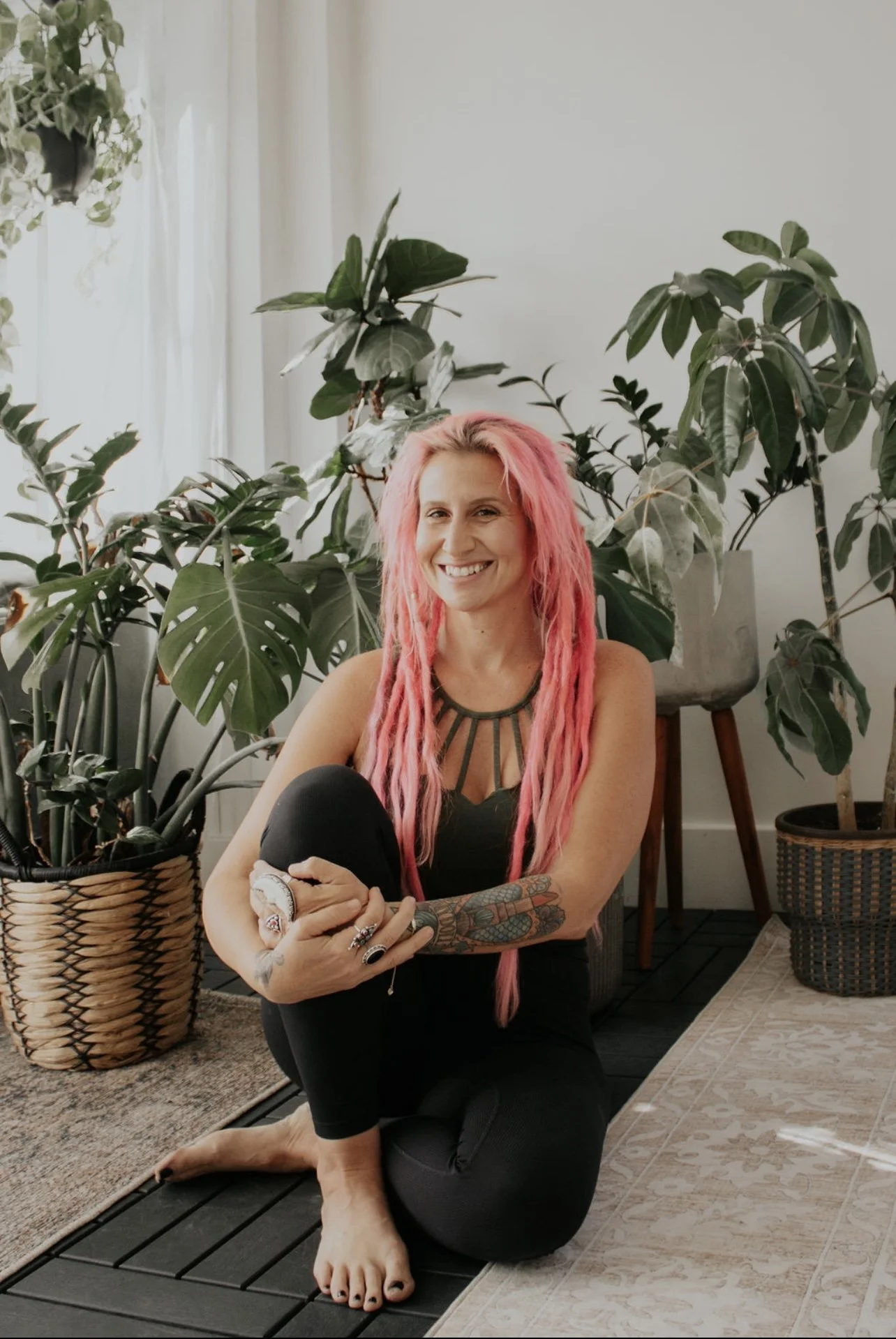 A woman with long pink dreadlocks sitting cross-legged on a mat indoors, surrounded by large potted plants, smiling at the camera.