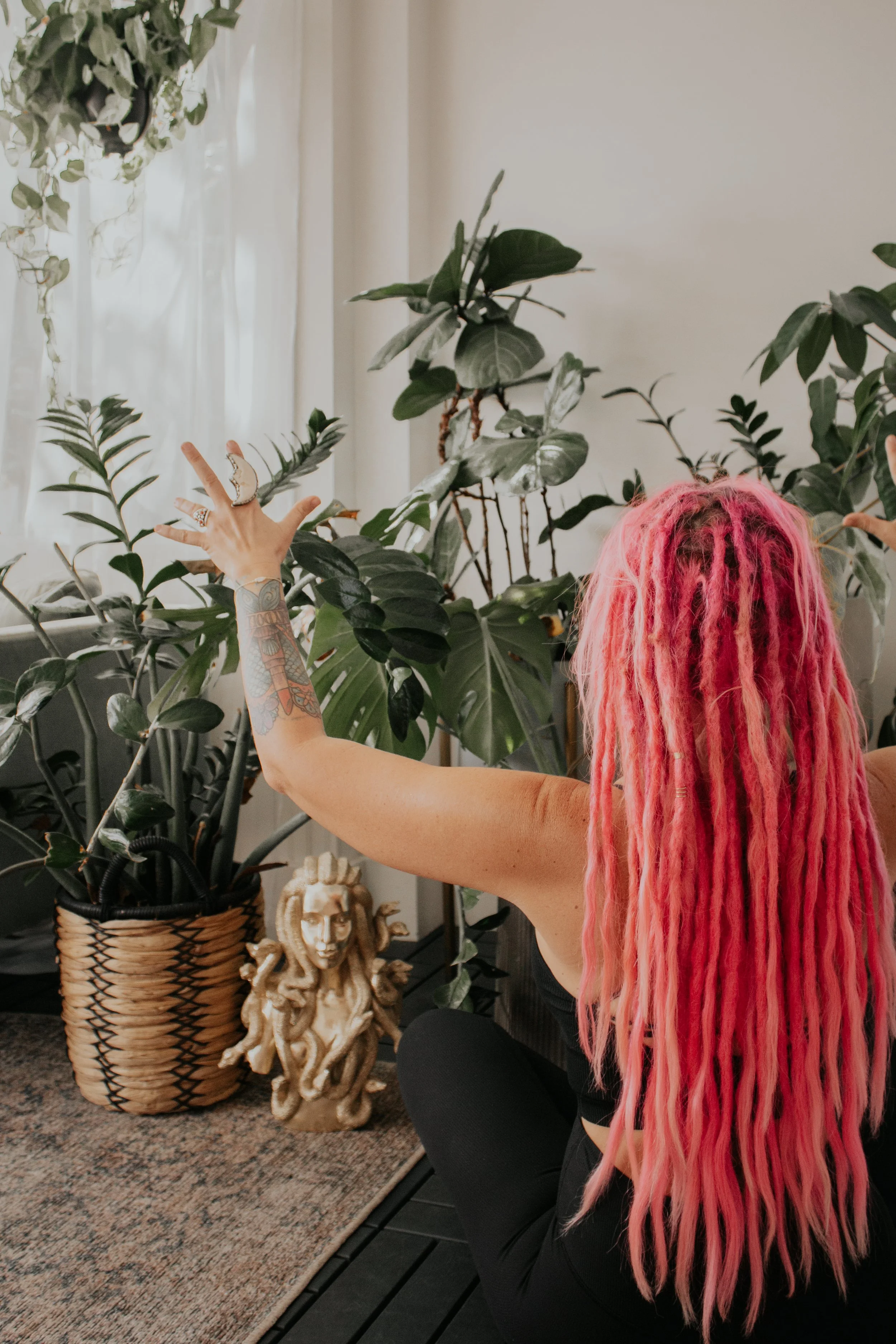 A woman with long pink dreadlocks and tattoos on her left arm, sitting in a room with plants, reaching out with her left hand. There is a decorative sculpture of Medusa on the floor, stocked with various potted plants, and a woven basket.