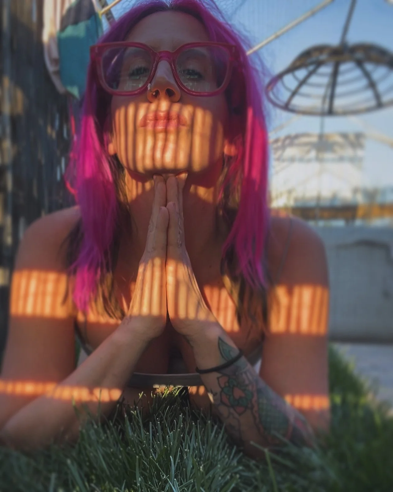 A woman with purple hair and glasses is posing outdoors with her hands in a prayer position. Shadows cast by a wooden structure create striped patterns on her face, arms, and chest. She is lying on grass with parasols and a fence in the background.