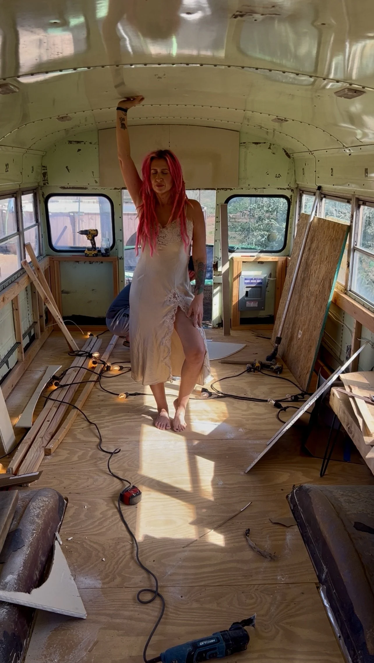 A woman with pink hair wearing a beige dress with lace trim standing barefoot inside an unfinished bus that is under renovation. She poses with one arm raised, touching the ceiling. The bus interior has exposed wooden framing, tools, and construction materials scattered around.